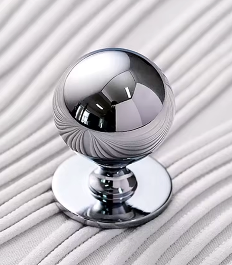 A polished chrome spherical furniture knob with a fluted lower section and a round base, shown on a textured white fabric background.