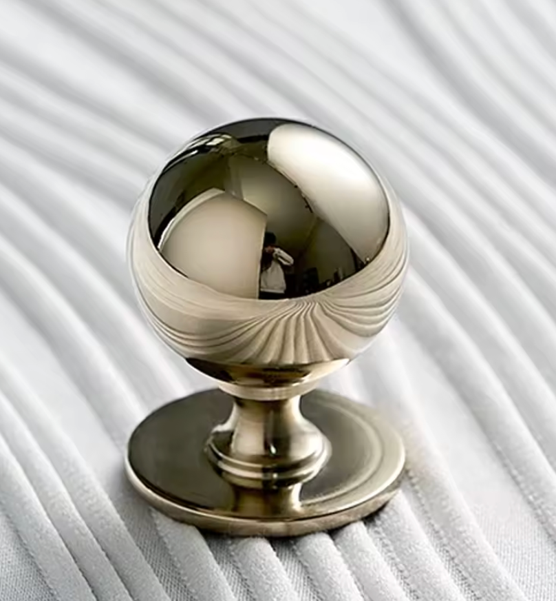A polished metallic cabinet knob with a spherical top and circular base, shown on a white ribbed fabric background.
