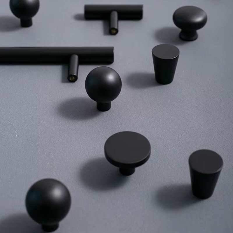 Matte black cabinet knobs in various shapes including round spheres, flat discs, and cylindrical forms, arranged on a textured gray surface.