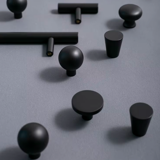 Matte black cabinet knobs in various shapes including round spheres, flat discs, and cylindrical forms, arranged on a textured gray surface.