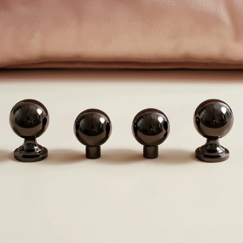 Four glossy black spherical cabinet knobs arranged in a row on a light beige surface, with a soft pink fabric or cushion blurred in the background.