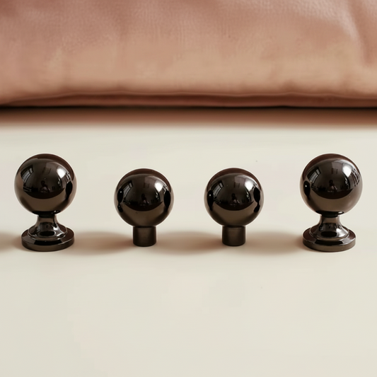 Four glossy black spherical cabinet knobs arranged in a row on a light beige surface, with a soft pink fabric or cushion blurred in the background.