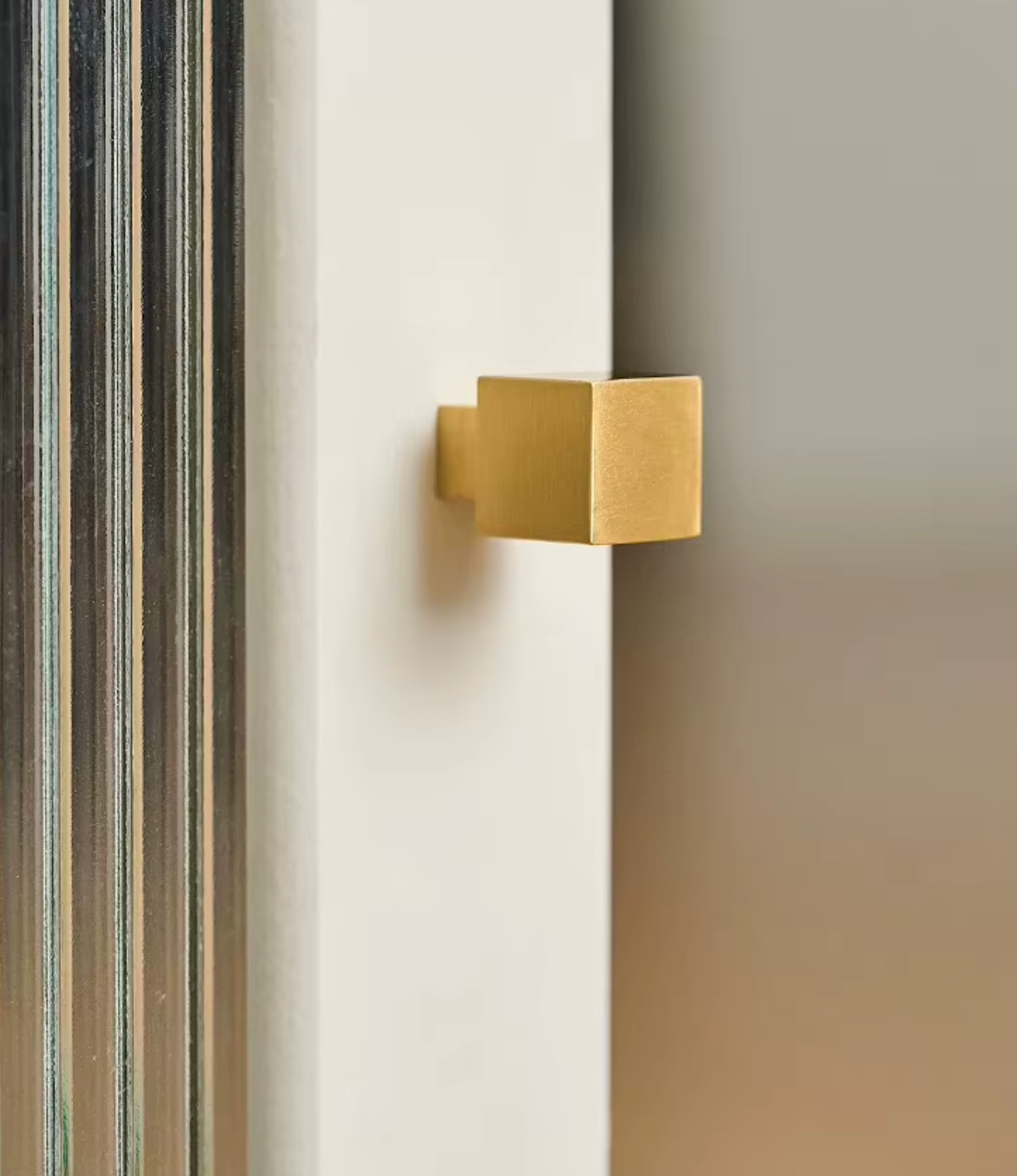 A square-shaped matte gold door knob mounted on a white door frame next to a glass pane with dark metal frames.