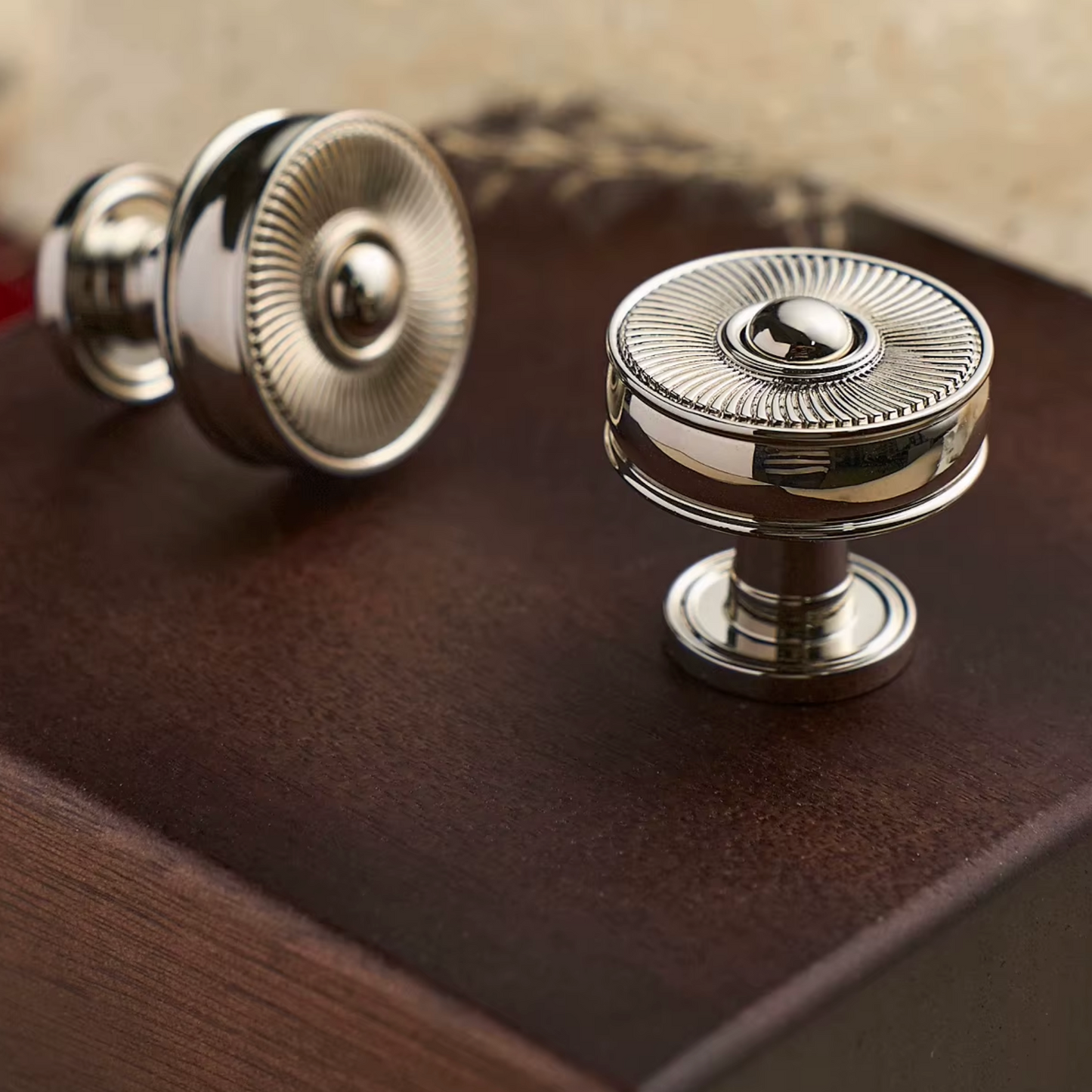 Two round polished metal cabinet knobs with a sunburst textured top and cylindrical base, displayed on a dark brown wooden surface.