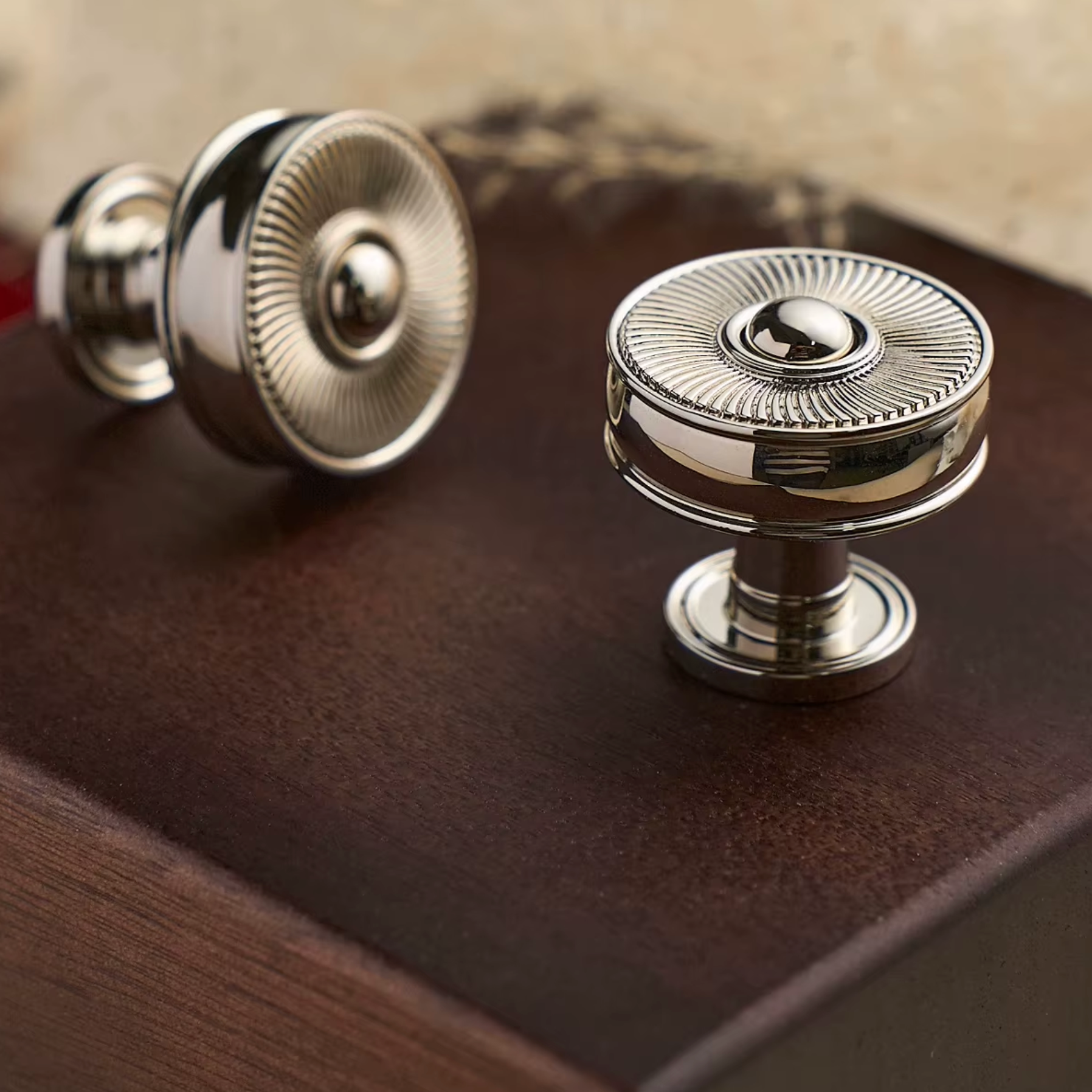 Two round polished metal cabinet knobs with a sunburst textured top and cylindrical base, displayed on a dark brown wooden surface.