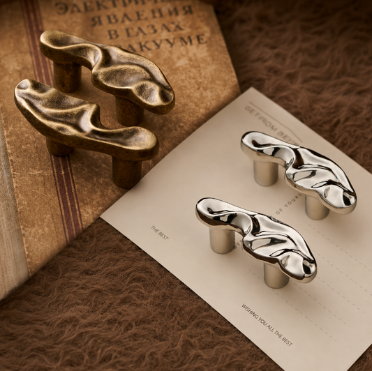 Two abstract sculptural cabinet knobs, one in polished silver and one in antique bronze, displayed on a styled flat-lay with a vintage book, a card, and a brown textured rug background.
