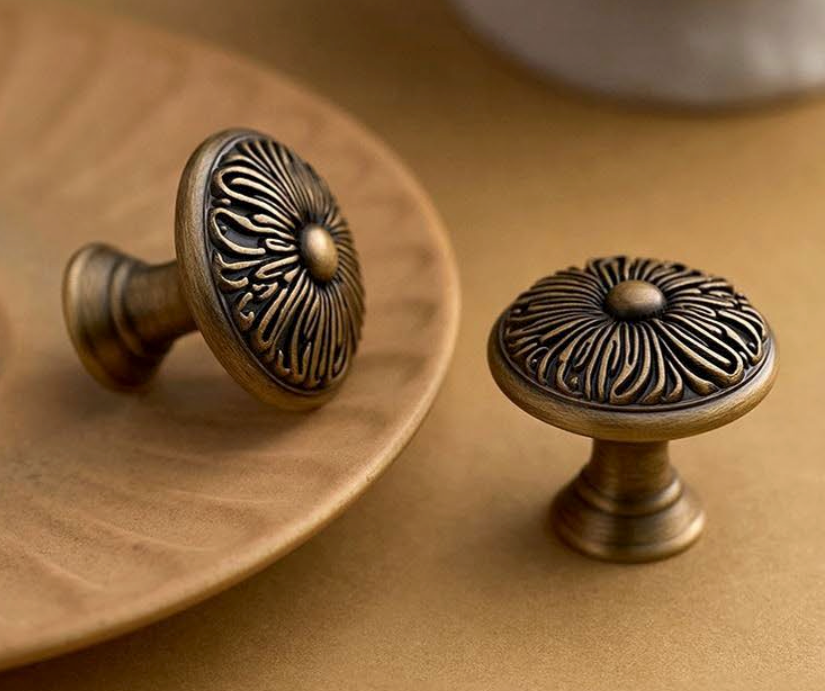 Two round antique bronze cabinet knobs with embossed floral patterns, shown on a warm brown surface with one resting on a wooden plate and the other standing beside it.