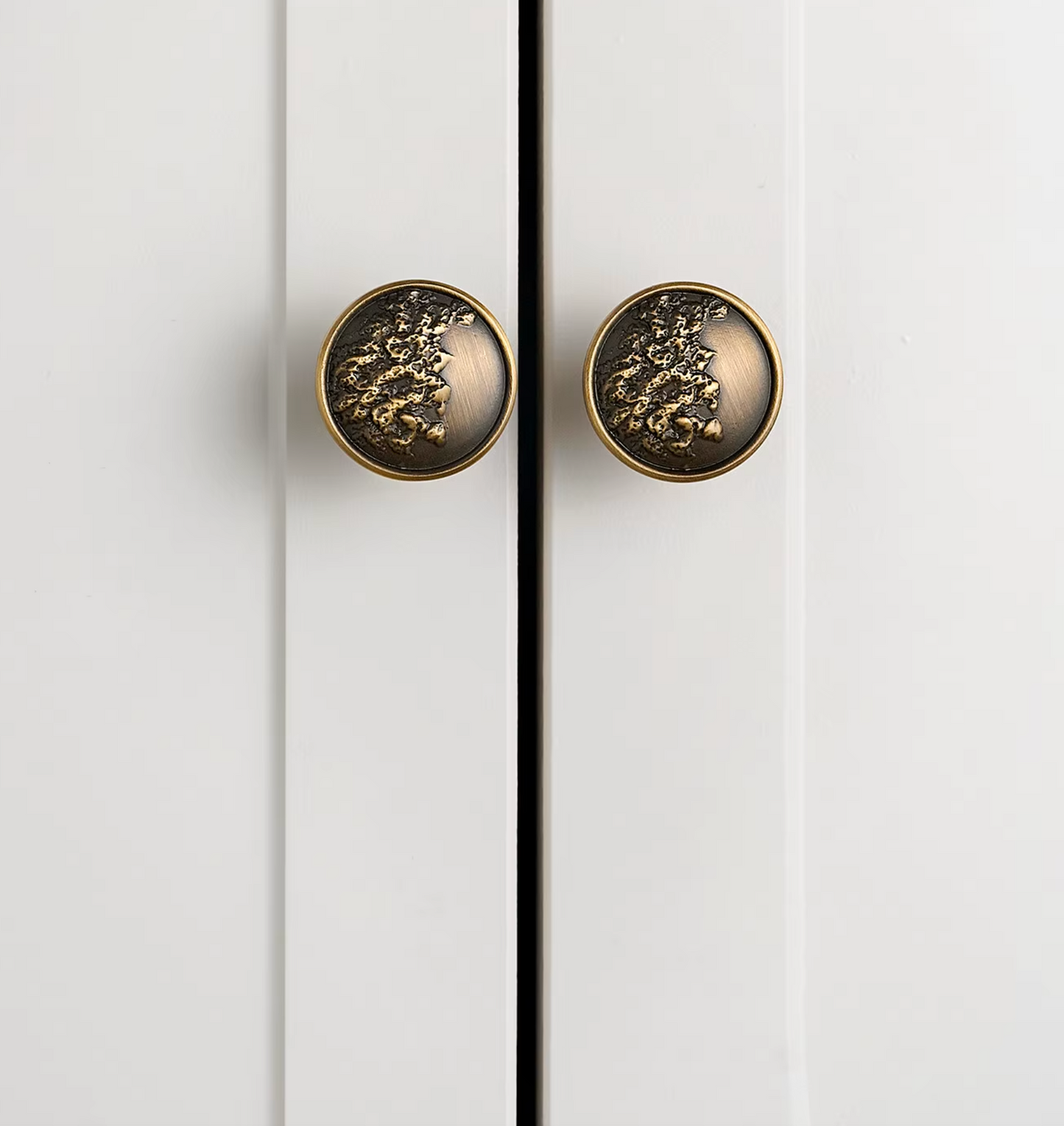 Two round cabinet knobs with a textured, metallic gold floral or leaf-like design on a dark bronze background, mounted on white paneled doors.