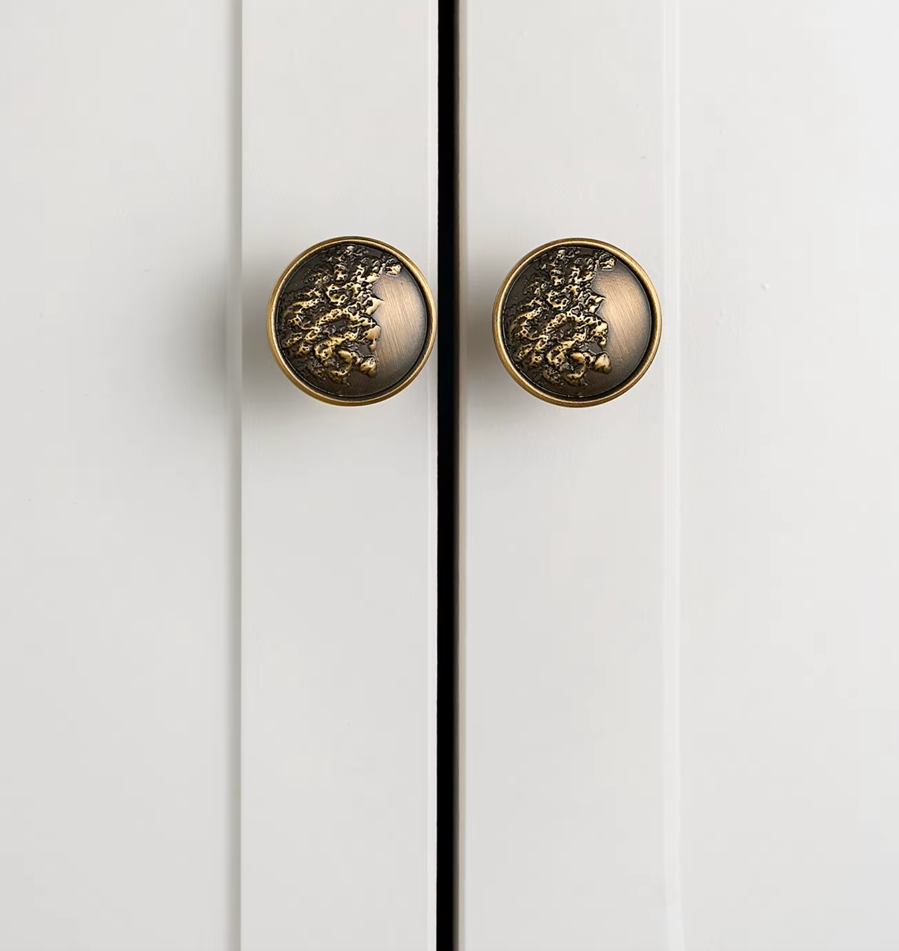 Two round cabinet knobs with a textured, metallic gold floral or leaf-like design on a dark bronze background, mounted on white paneled doors.
