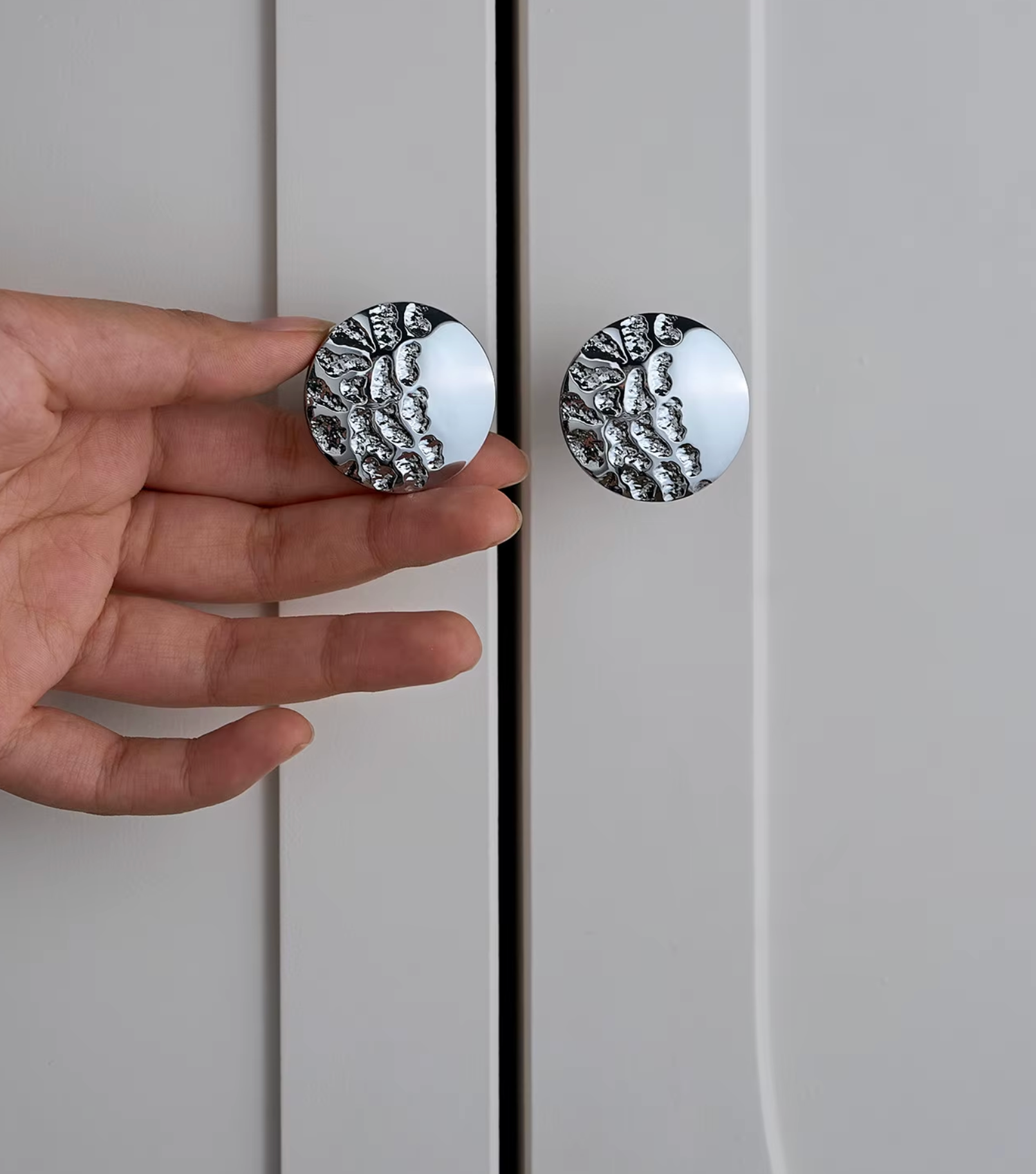 A hand holds a round silver cabinet knob with a textured, organic pattern against a light gray door, next to another matching knob.