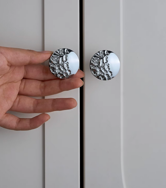 A hand holds a round silver cabinet knob with a textured, organic pattern against a light gray door, next to another matching knob.
