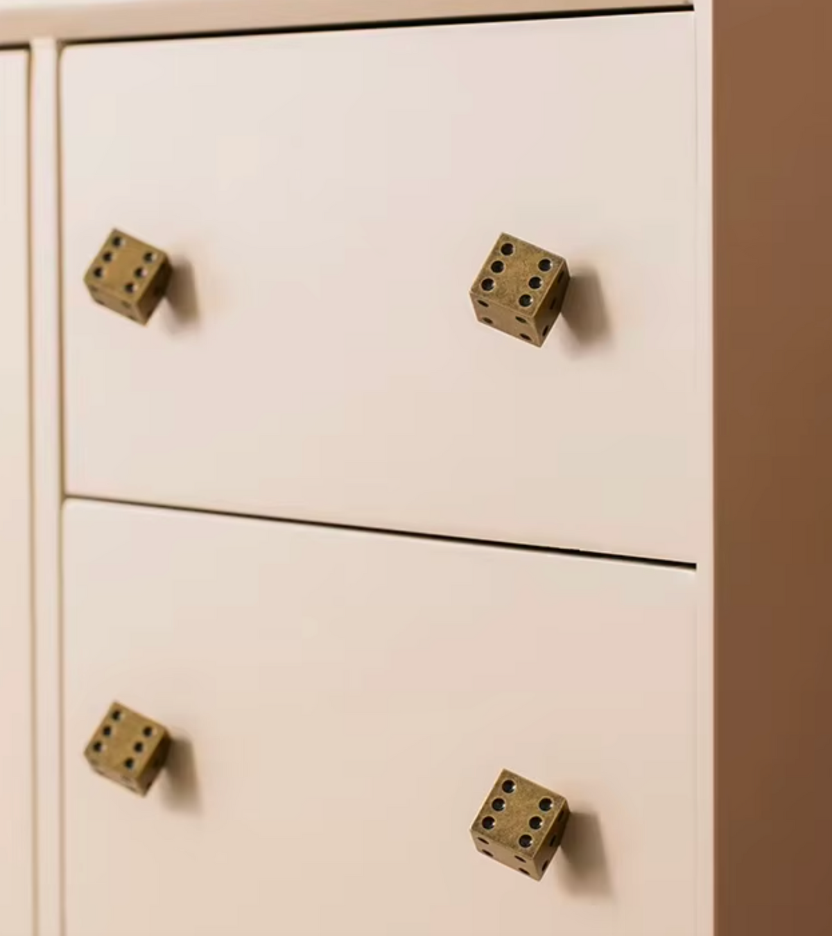 A close-up of a cream-colored dresser with four metallic bronze dice-shaped drawer pulls.