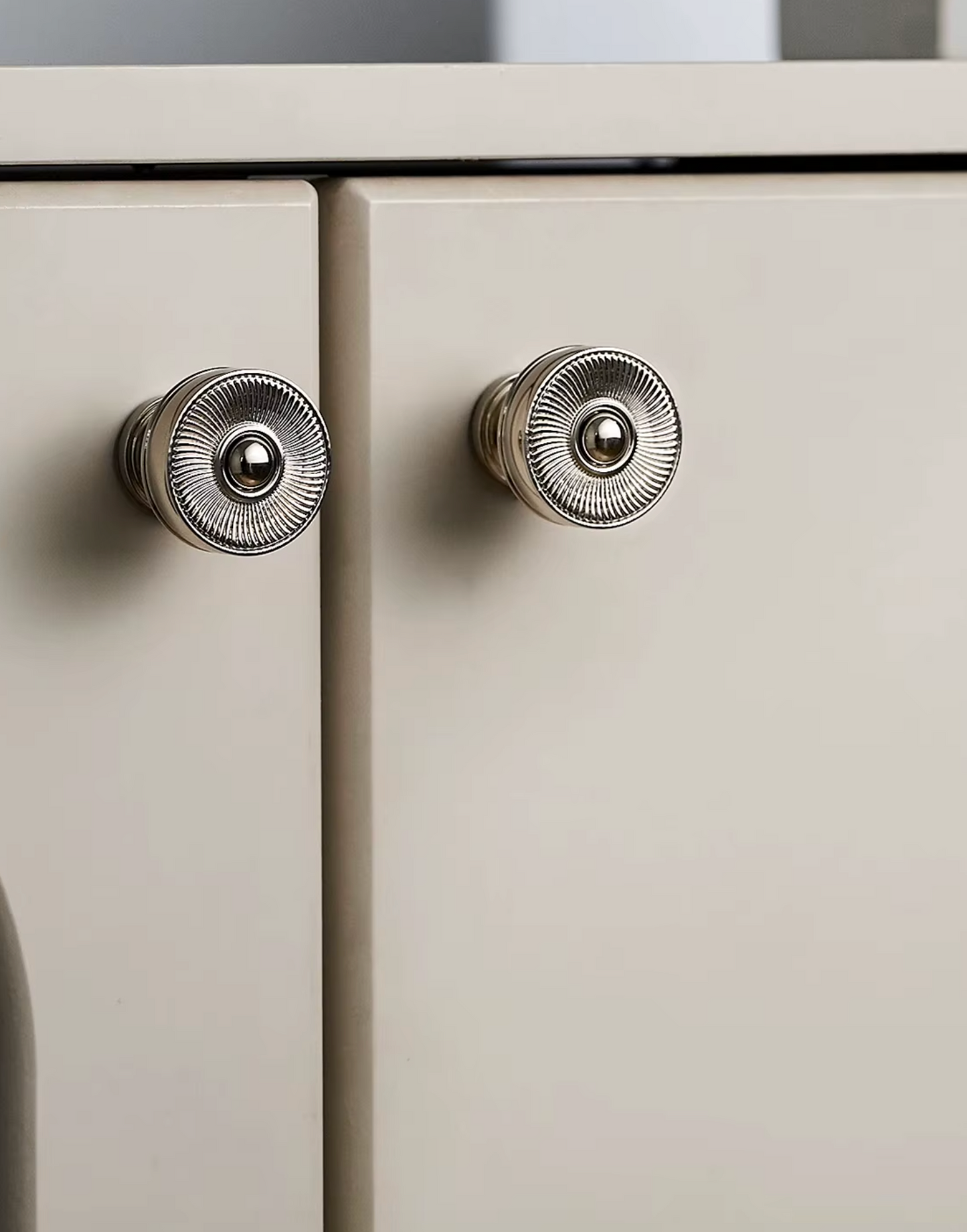 Two round silver metal cabinet knobs with a ribbed textured pattern, mounted on light beige doors.