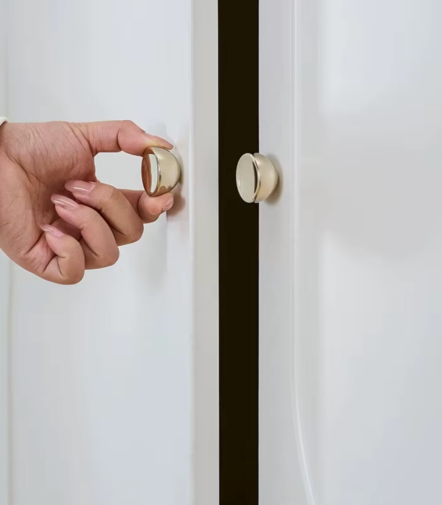 A hand holding a round, brushed brass door stopper against a white door, with a matching door stopper installed on the adjacent door.