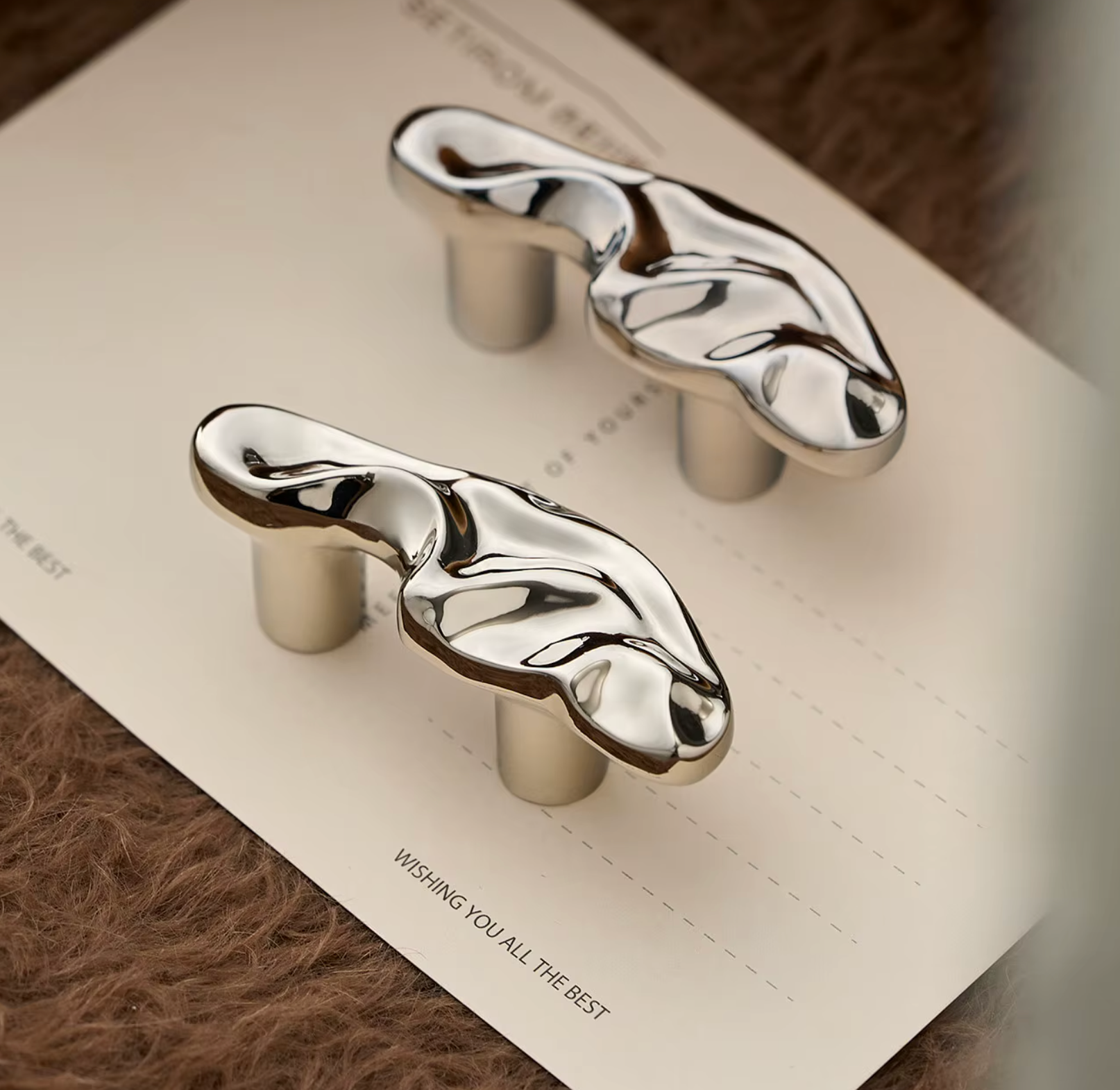 Two polished chrome cabinet knobs with an organic, fluid wavy shape, mounted on a white card over a brown textured surface.
