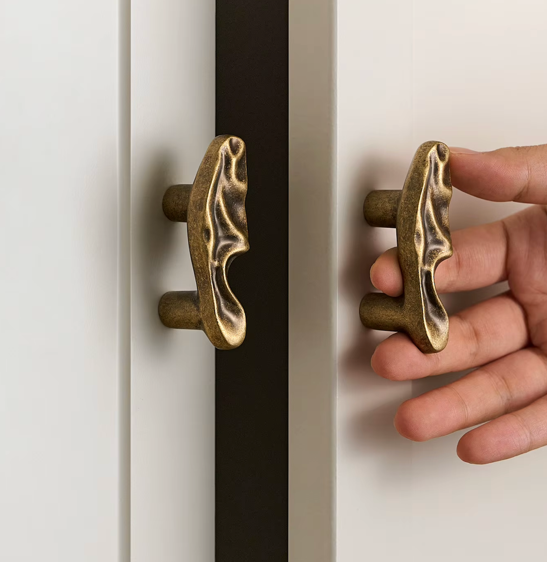 A close-up of a hand holding a distressed bronze cabinet pull handle next to a white door, with another matching handle visible on a dark door panel.