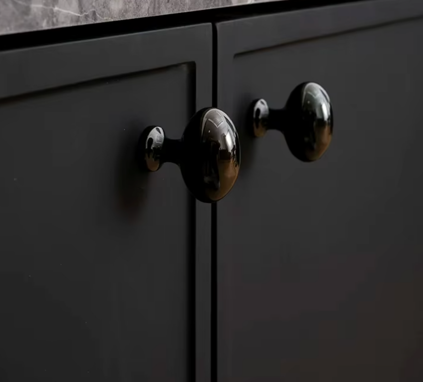 Close-up of glossy black oval cabinet knobs mounted on matte black cabinet doors, with a dark stone countertop visible at the top.