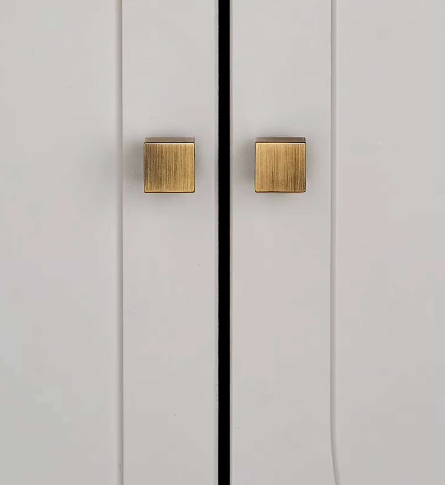 Two square brushed brass cabinet knobs mounted on white paneled doors with a black center line.