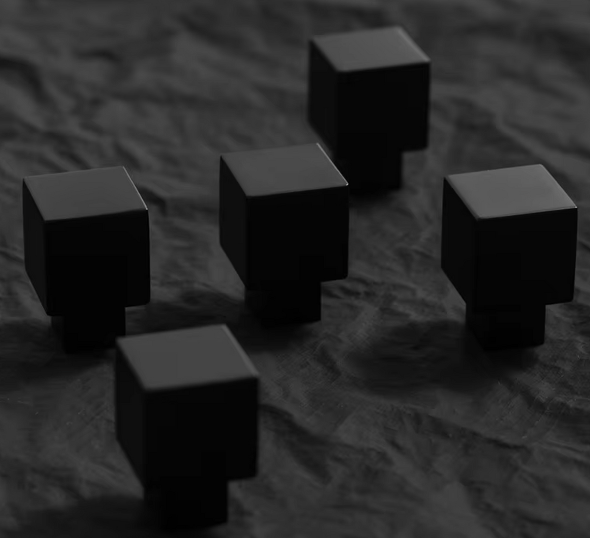 Five matte black cube-shaped objects arranged on a dark textured fabric surface.