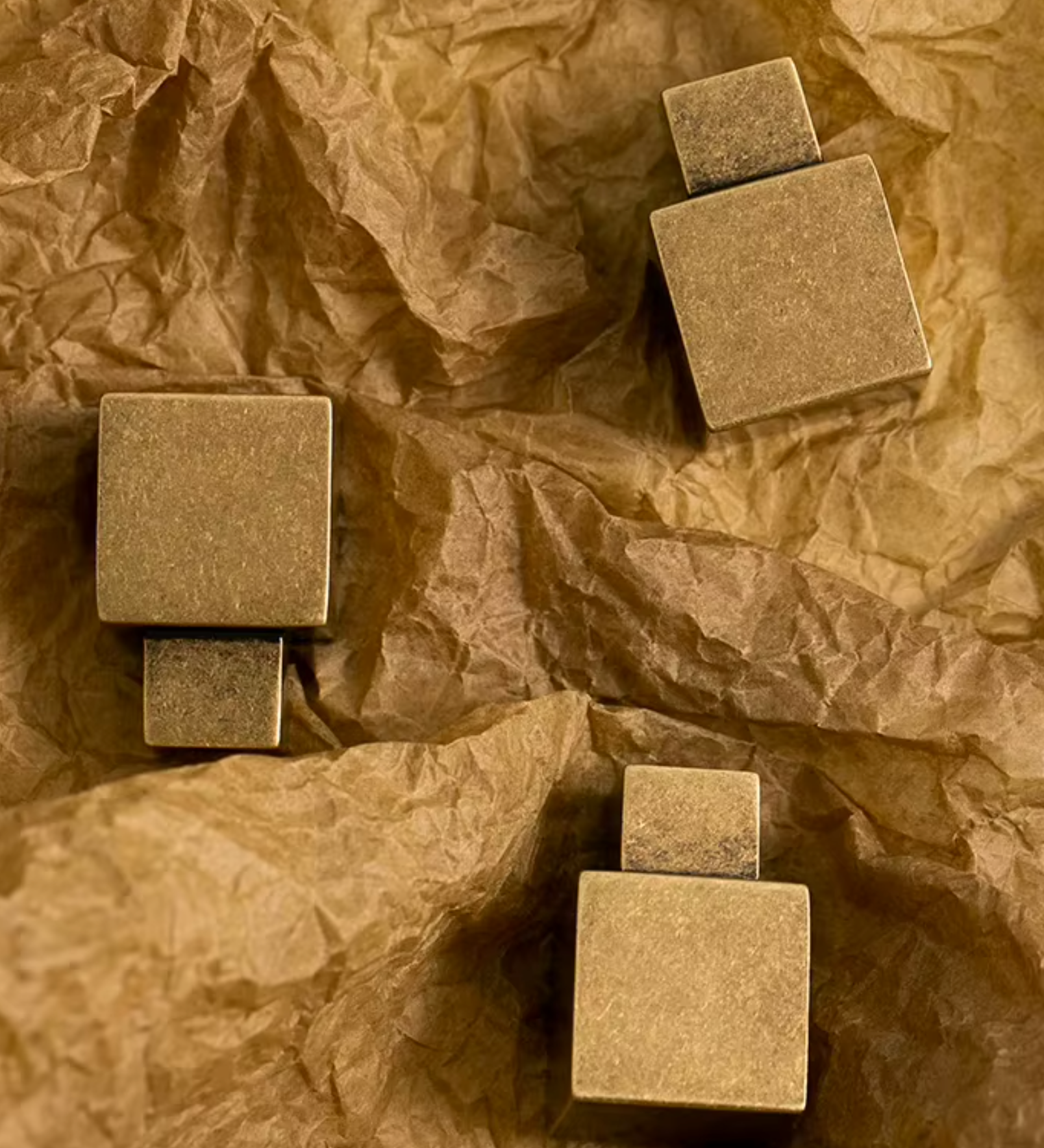 Three square stone-like jewelry pieces with a textured matte finish, arranged on crumpled brown paper.
