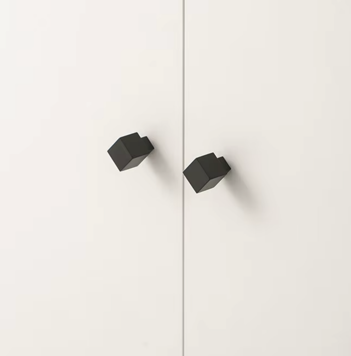 Two matte black geometric cabinet knobs mounted on a white door, shown from an overhead angle with a thin vertical seam in the background.