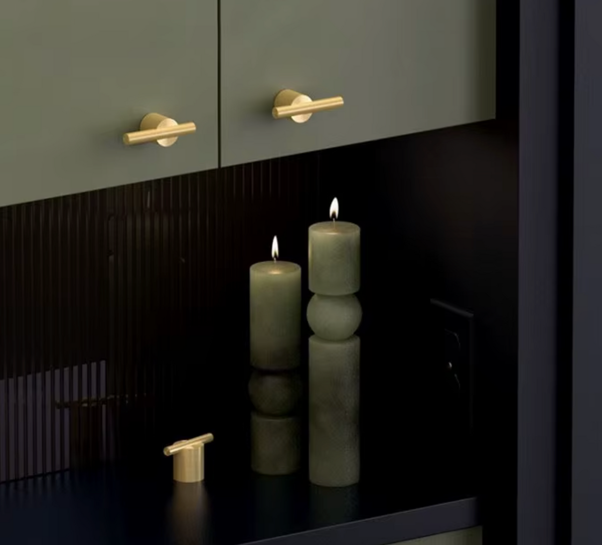 Modern minimalist hardware set featuring matte black cabinets with gold bar handles, a matching gold wall-mounted fixture, and two textured sage green pillar candles on a dark reflective surface.