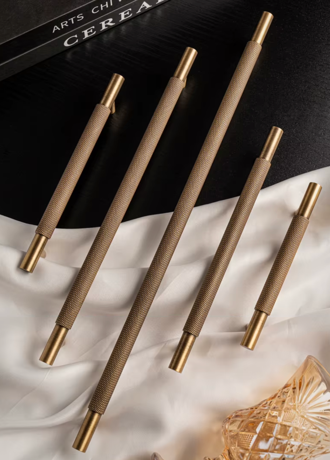 Five modern cabinet pulls with textured champagne-gold handles and polished brass ends, arranged diagonally on a black and white draped fabric background.