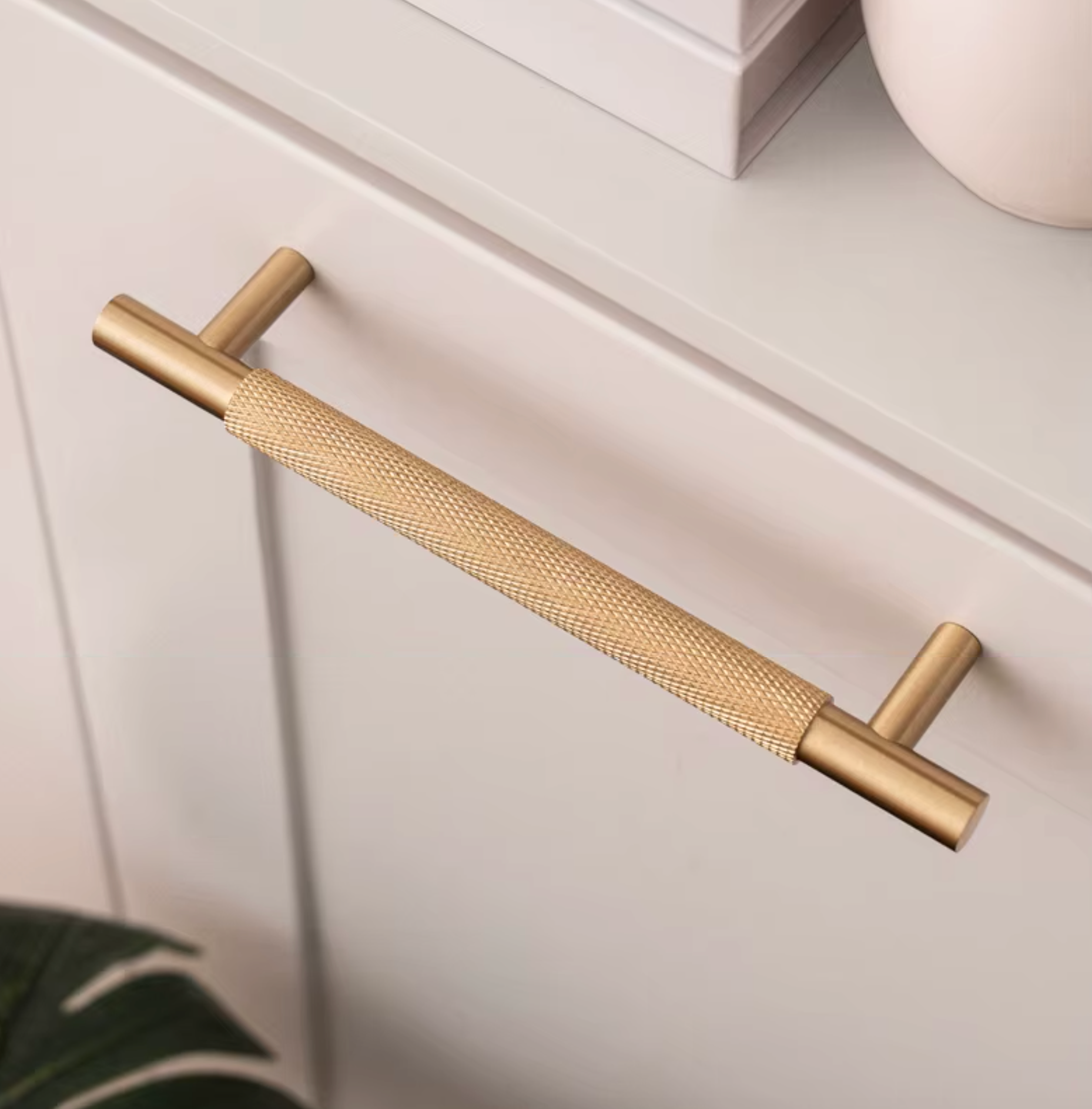 A modern two-piece cabinet pull handle in brushed gold and textured brass, mounted on a white drawer front.