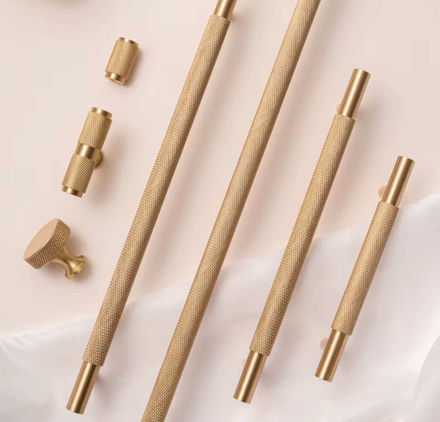 A set of brushed brass cabinet hardware, including three long knurled bar pulls, two matching knob pulls, and two smaller cylindrical pulls, arranged on a light beige background with a soft white fabric drape.