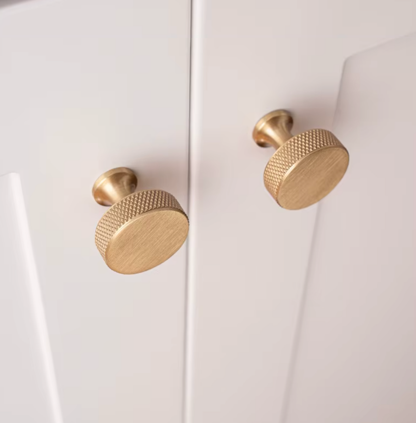 Two round brushed brass cabinet knobs with a knurled textured edge, mounted on a white paneled cabinet door.