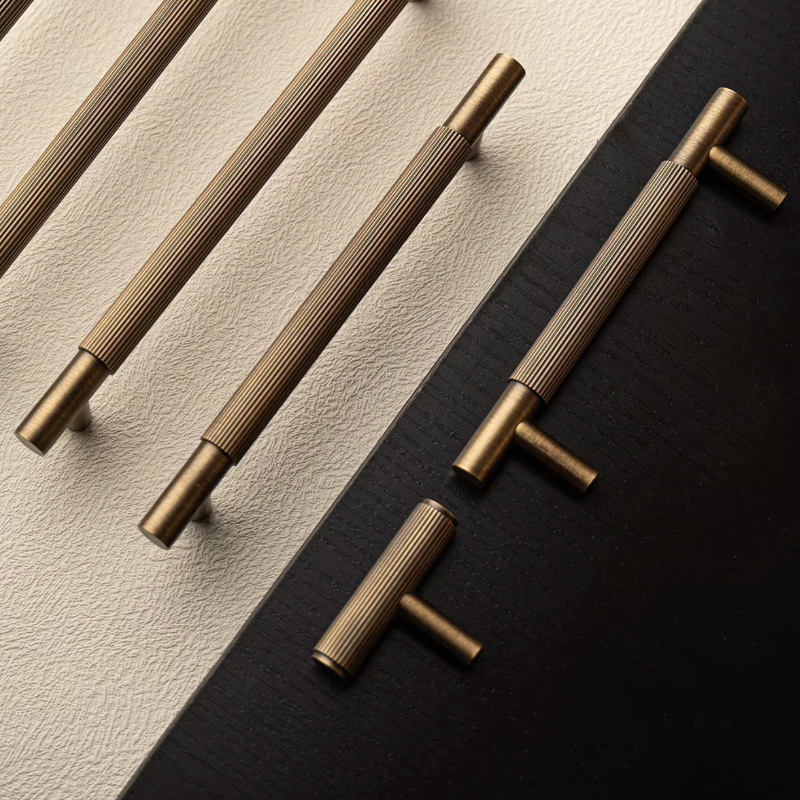 Four elongated cabinet pulls and two T-bar cabinet pulls in a brushed antique brass finish, arranged diagonally on a split beige and black background.