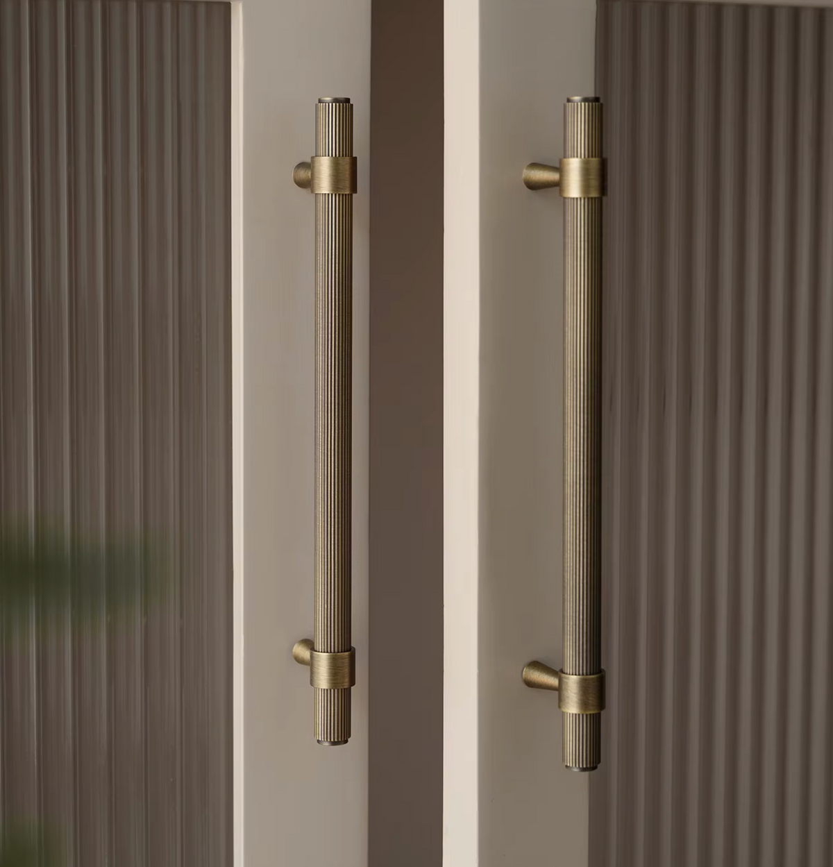 Two long vertical brushed brass door handles mounted on a beige door with ribbed brown panels on either side.