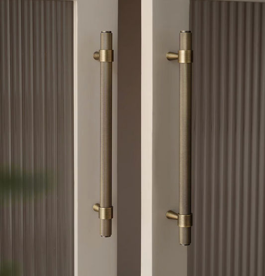 Two long vertical brushed brass door handles mounted on a beige door with ribbed brown panels on either side.