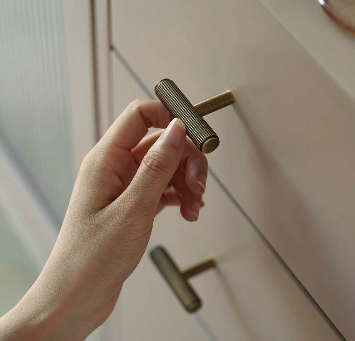 A close-up of a hand holding a cylindrical ribbed metal cabinet knob against a light beige drawer front.