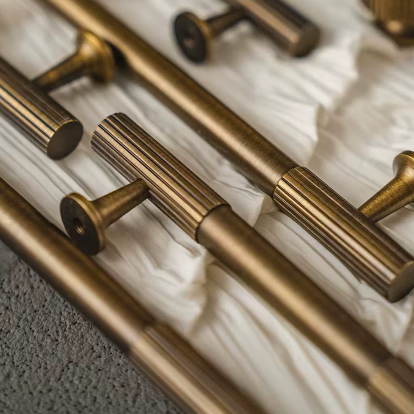 Close-up of multiple brass-toned wall or cabinet handles arranged diagonally on white tissue paper, with a textured gray surface visible at the lower left.