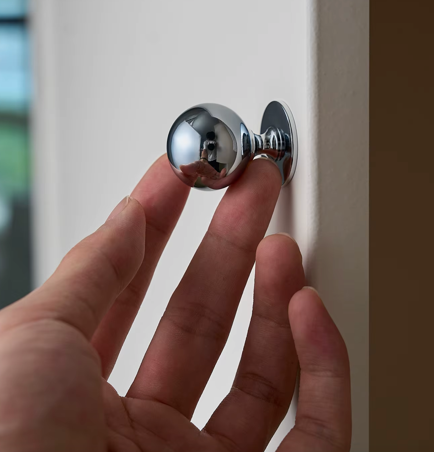 A hand holding a small, polished chrome doorknob in front of a plain off-white wall.