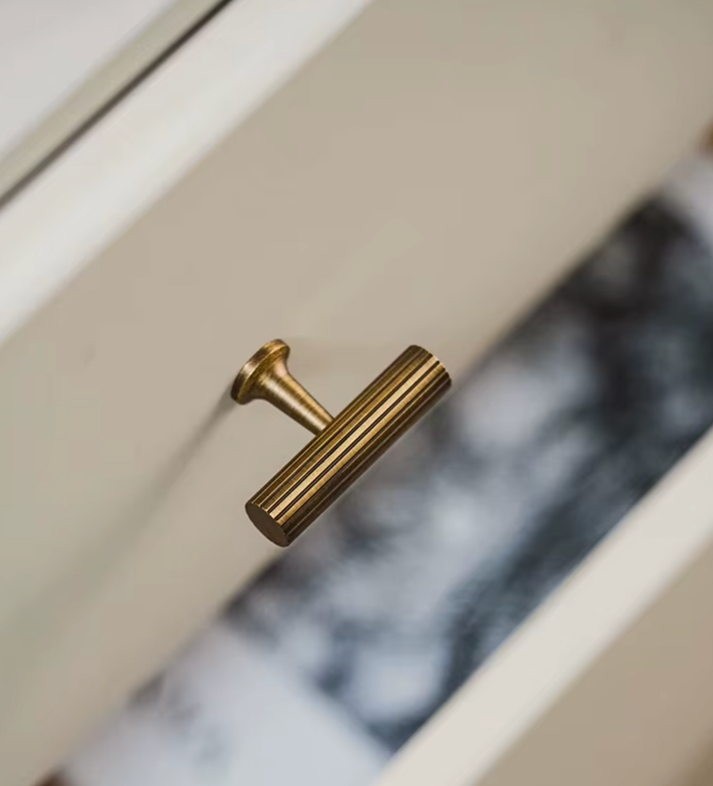 A close-up of a cylindrical brushed brass cabinet pull handle mounted on a white drawer front, with a blurred interior background visible behind it.