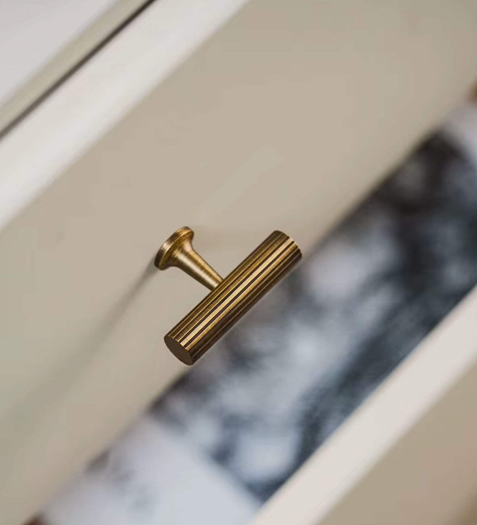 A close-up of a cylindrical brushed brass cabinet pull handle mounted on a white drawer front, with a blurred interior background visible behind it.
