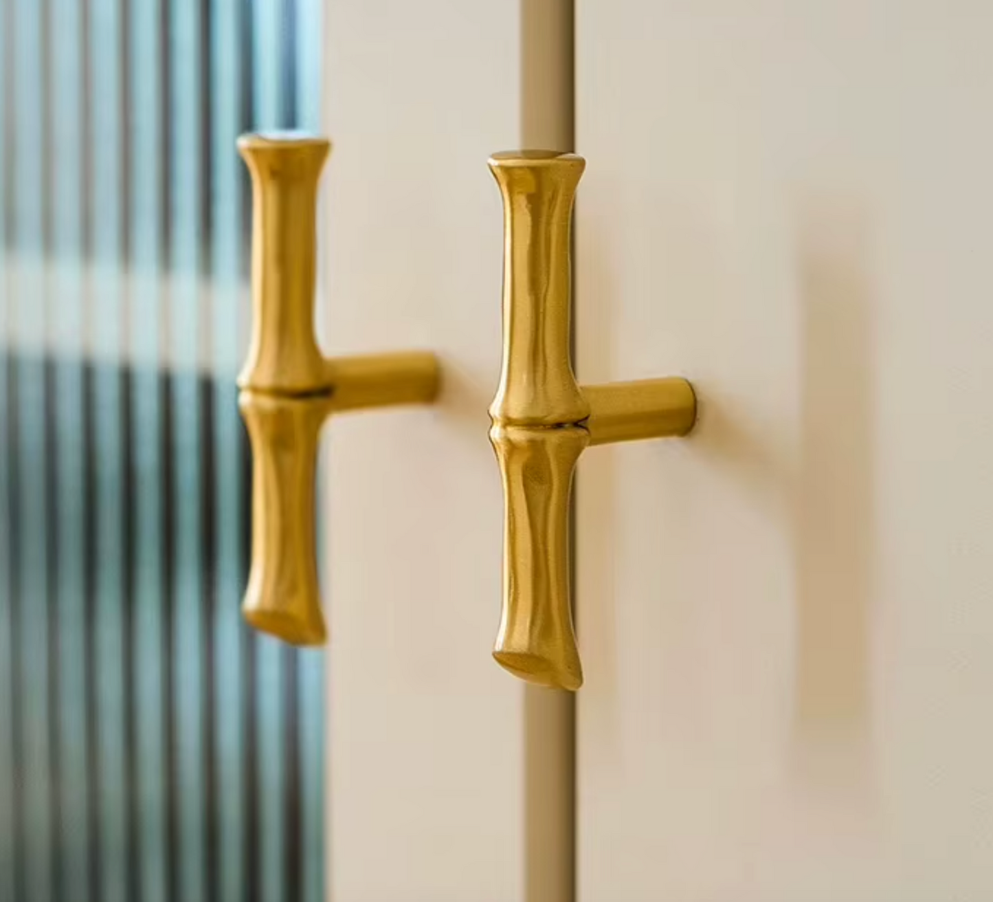 Two gold bamboo-style door handles mounted on a cream-colored door, with a softly blurred striped window or glass panel in the background.