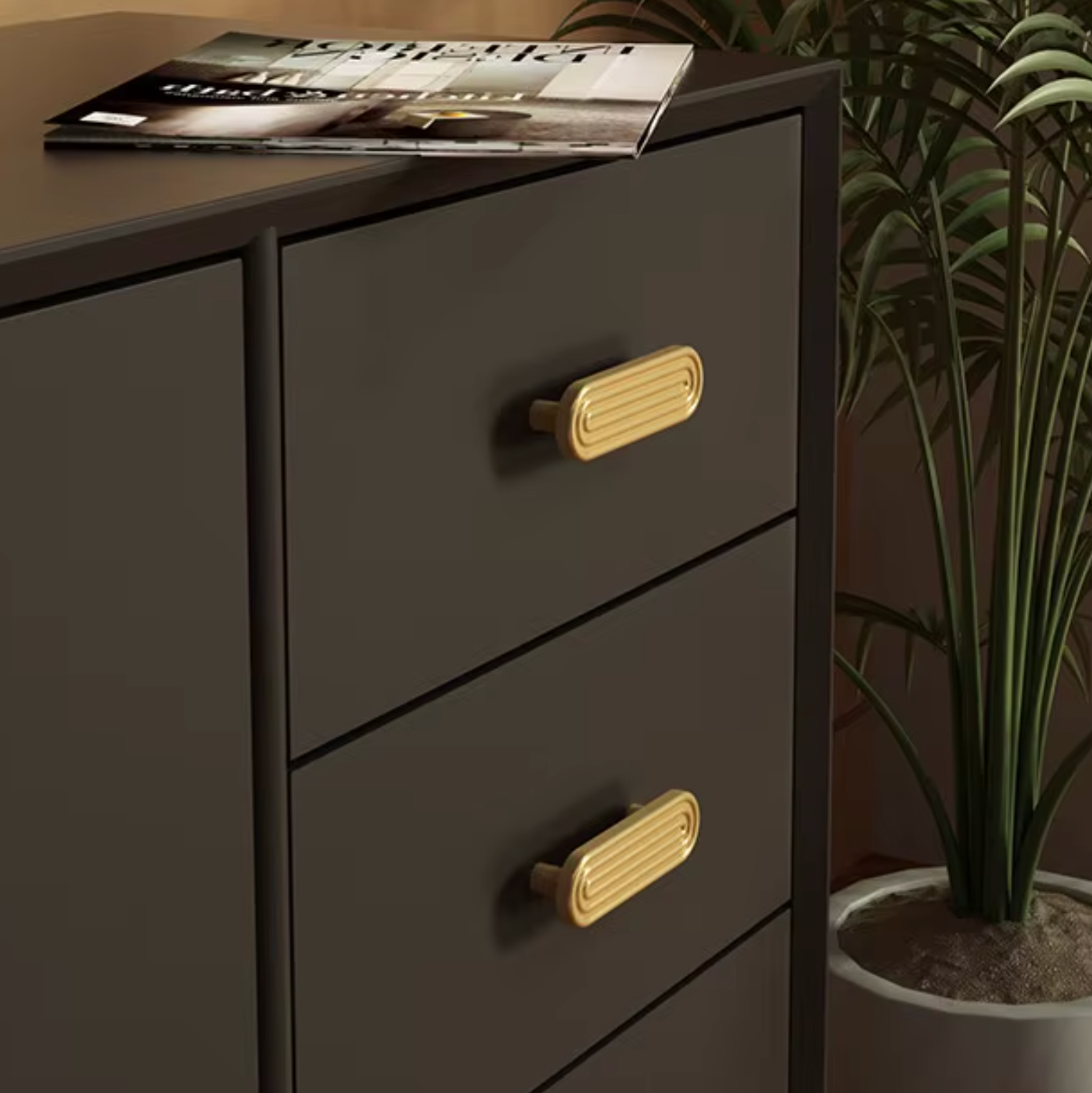A dark charcoal gray three-drawer dresser with ribbed gold metal handles, shown next to a potted plant and with a magazine on top.