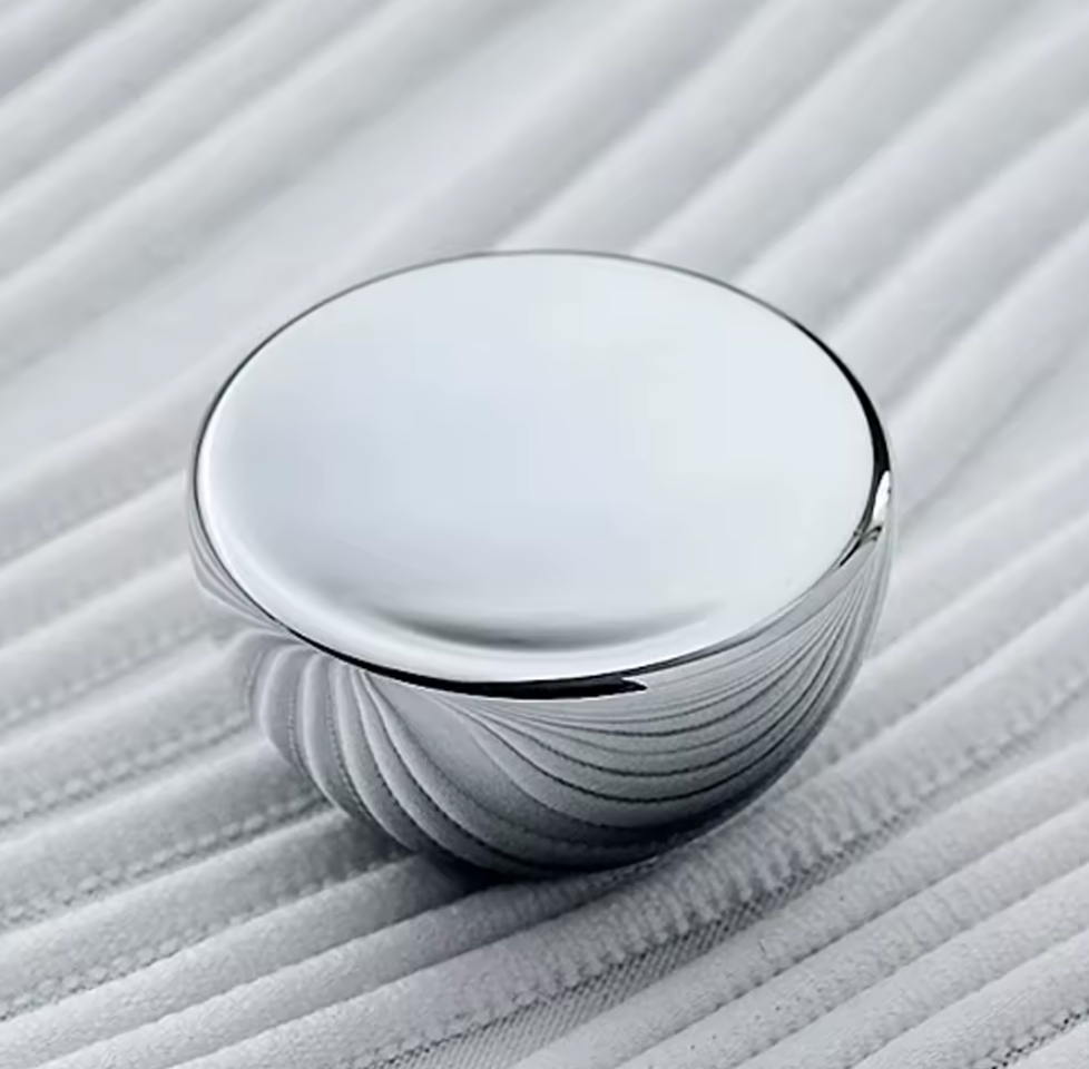 A round, polished silver metal knob with a smooth top surface and a ribbed textured base, resting on a white ribbed fabric surface.