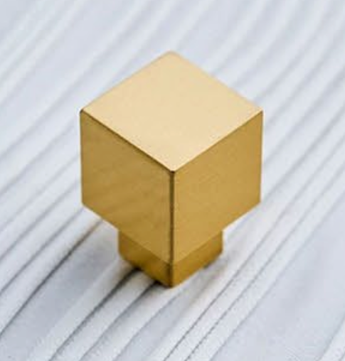A square gold cabinet pull knob with a brushed metallic finish, resting on a white ribbed surface.