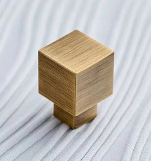 A square-shaped brass cabinet knob with a brushed finish, standing upright on a ribbed white fabric background.